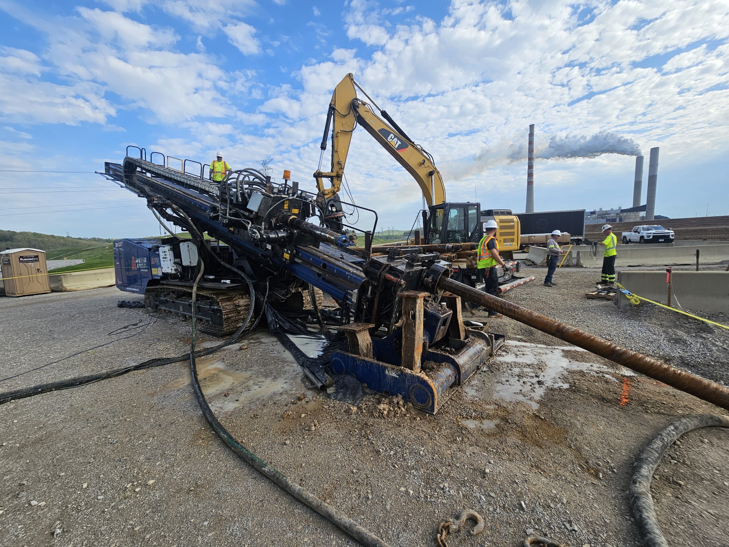 Horizontal drilling rig and workers at coal ash dewatering site, with heavy machinery and power plant in background, illustrating Ellingson-DTD's innovative dewatering technology.