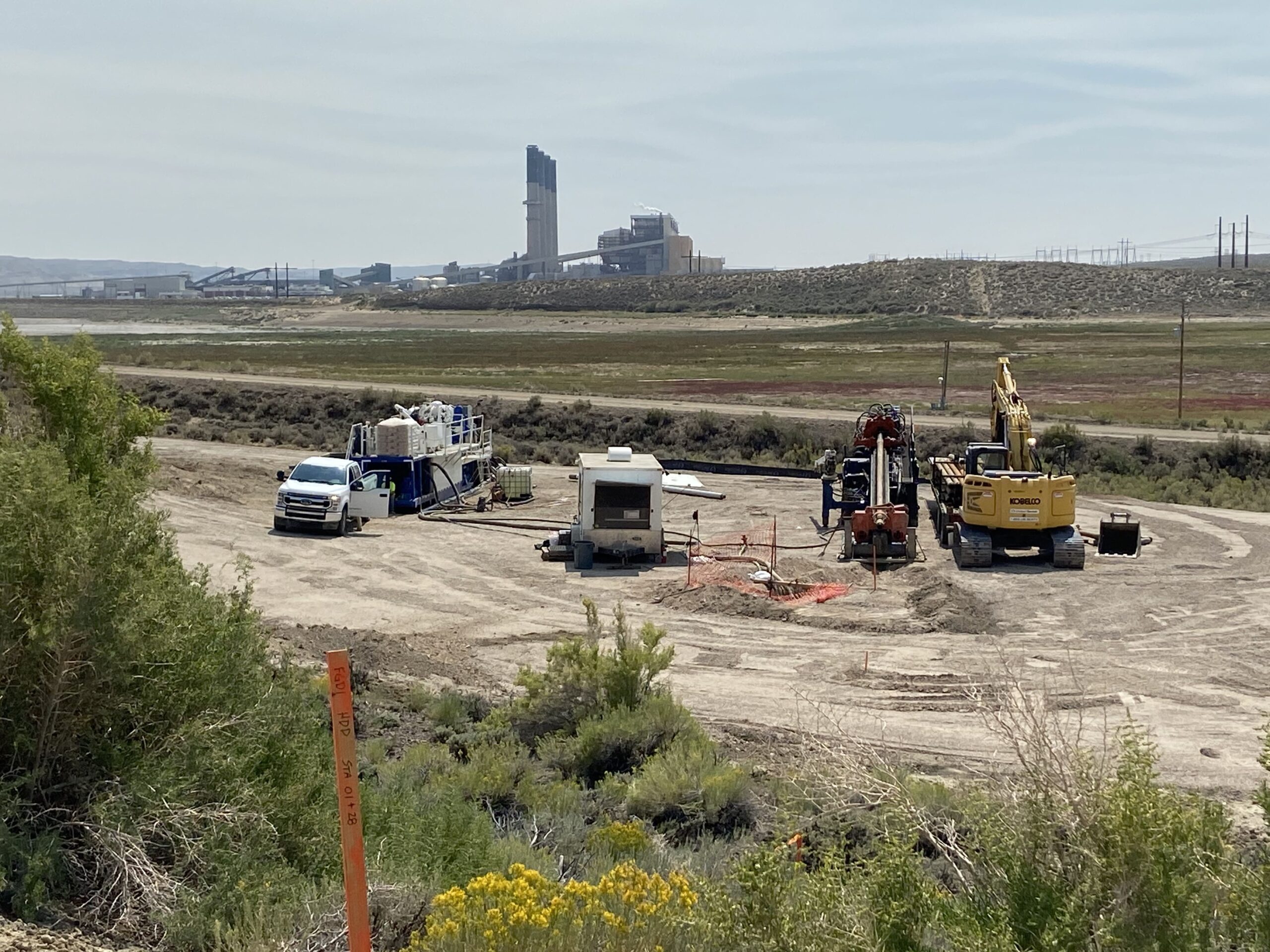 Construction site for groundwater containment project with drilling equipment, trucks, and flue gas desulfurization plant in background.