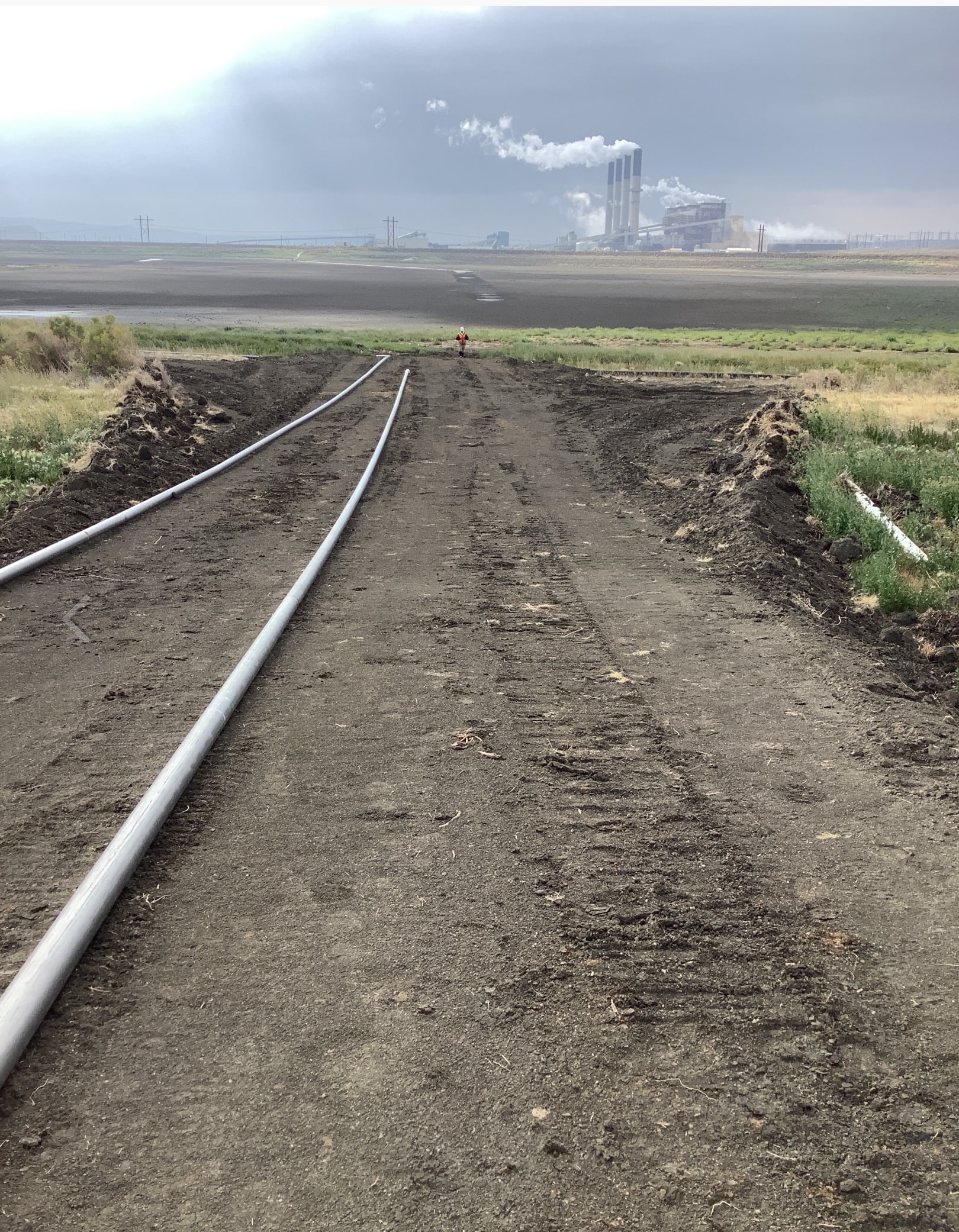 Dirt road with two horizontal pipes leading towards a coal power plant in the background, illustrating Ellingson Companies' groundwater containment and coal ash dewatering project.